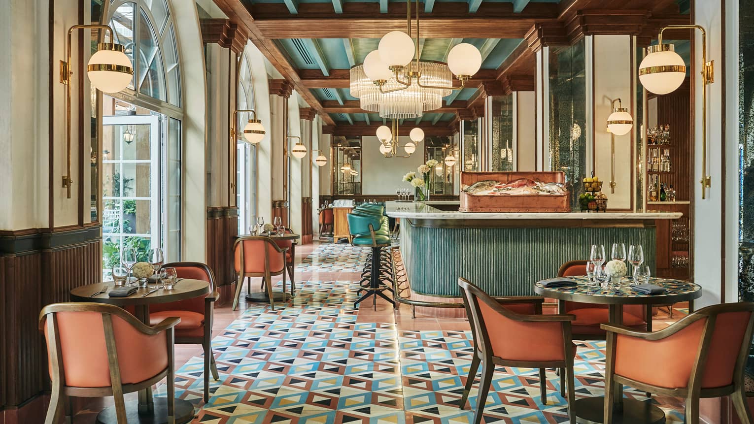 Interior of a restaurant dining room with patterned floor in blue, black, white and coral colours, coral-and-wood dining chairs, round wooden tables and a blue-and-white counter in the center