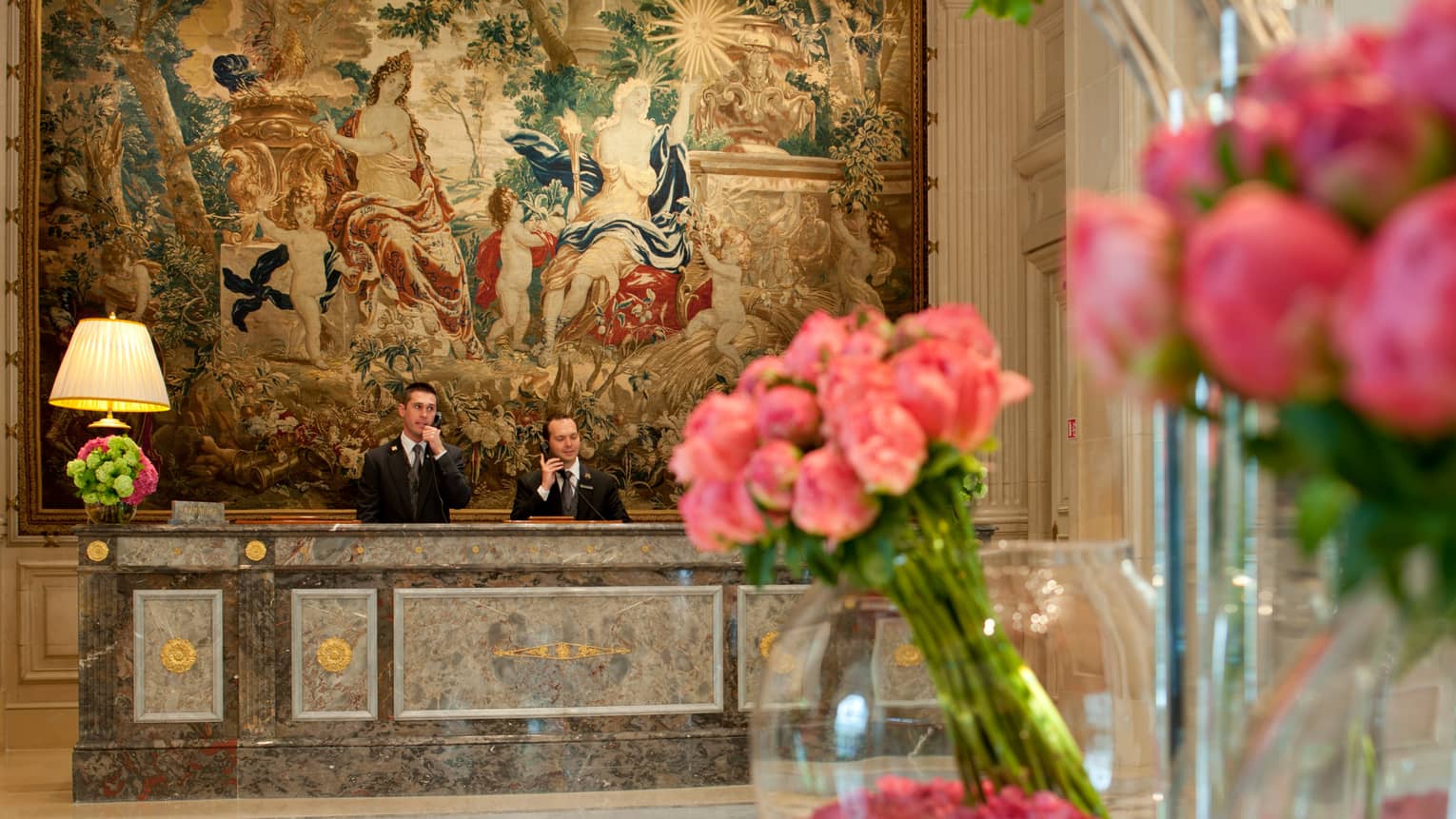 Hotel staff answer telephones at marble reception desk under large mural past pink flowers