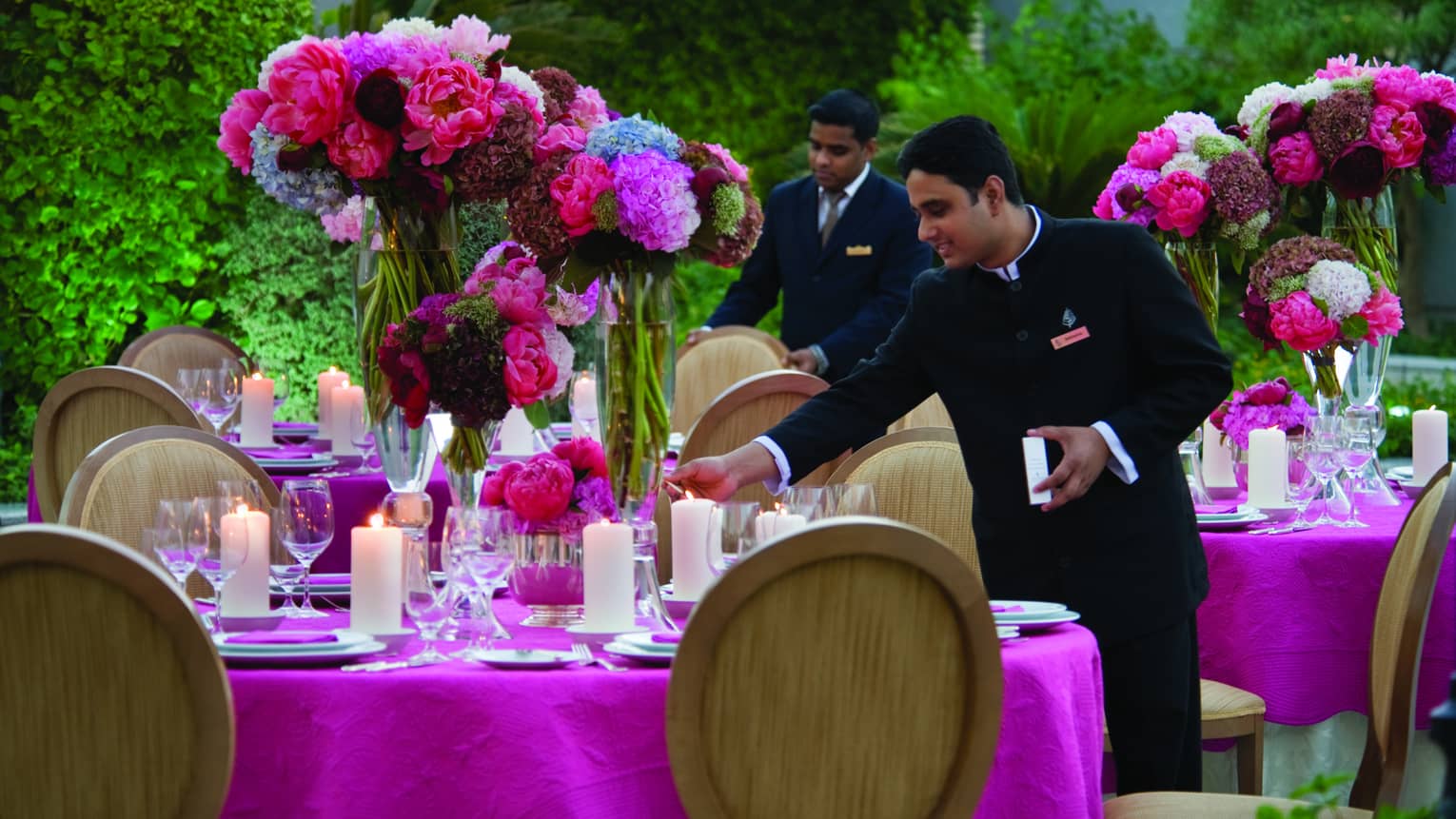 Two male staff set table with pink tablecloth, large vases of flowers and candles