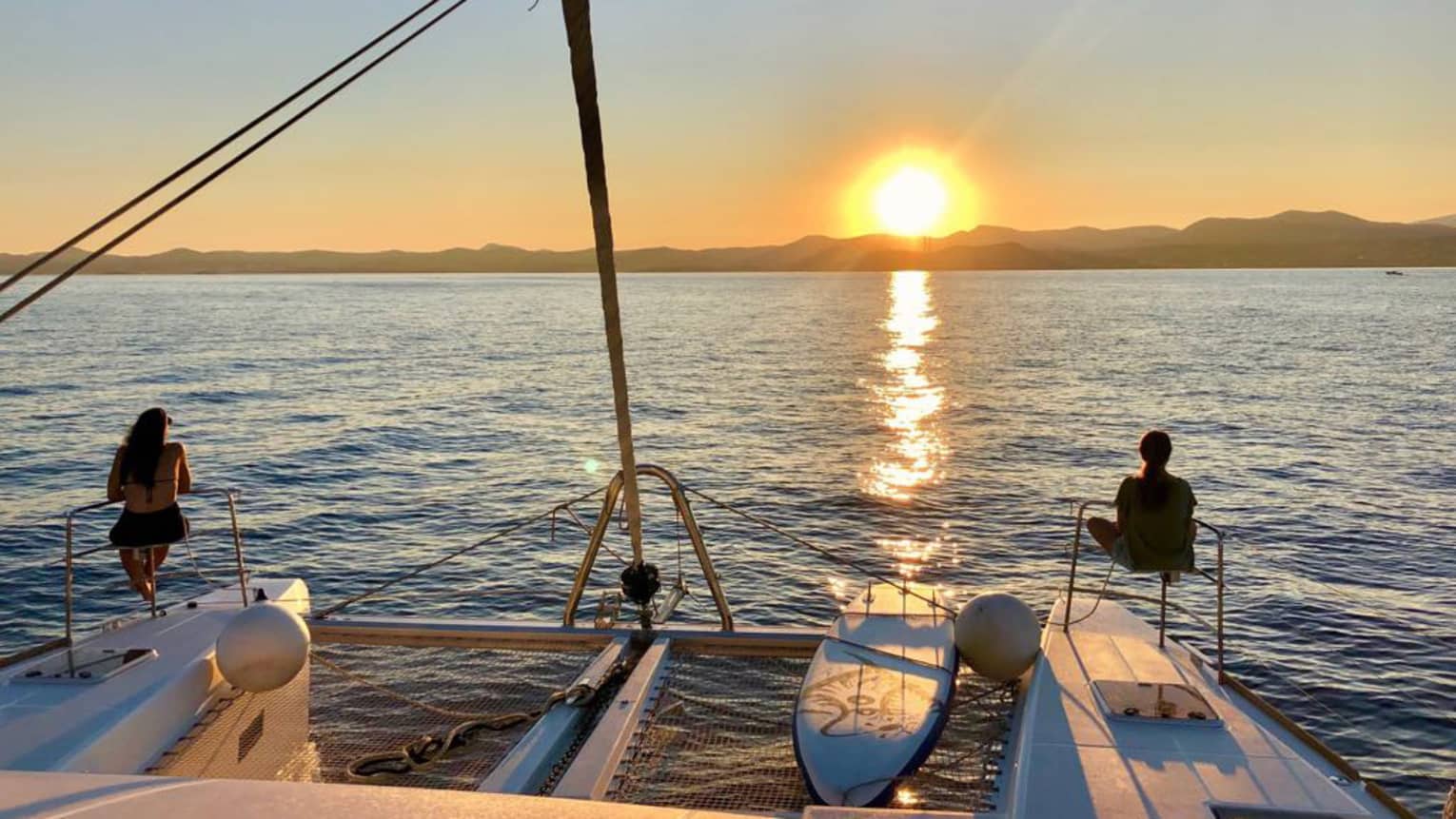 Two guests lounge on luxury sailboat near stern railing at sunset