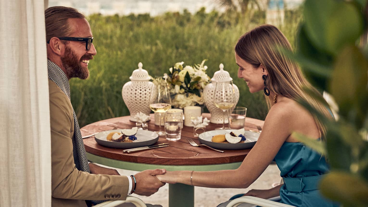 Smiling couple holds hands at dinner set with fruit plates, wine, flowers and two white perforated porcelain candle holders.