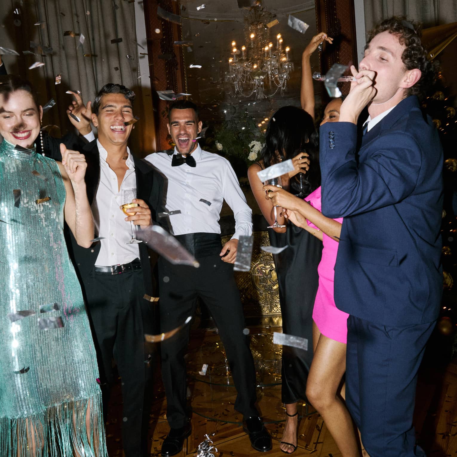 Six people dressed in cocktail party attire smile and celebrate as metallic confetti falls over them in a hotel suite