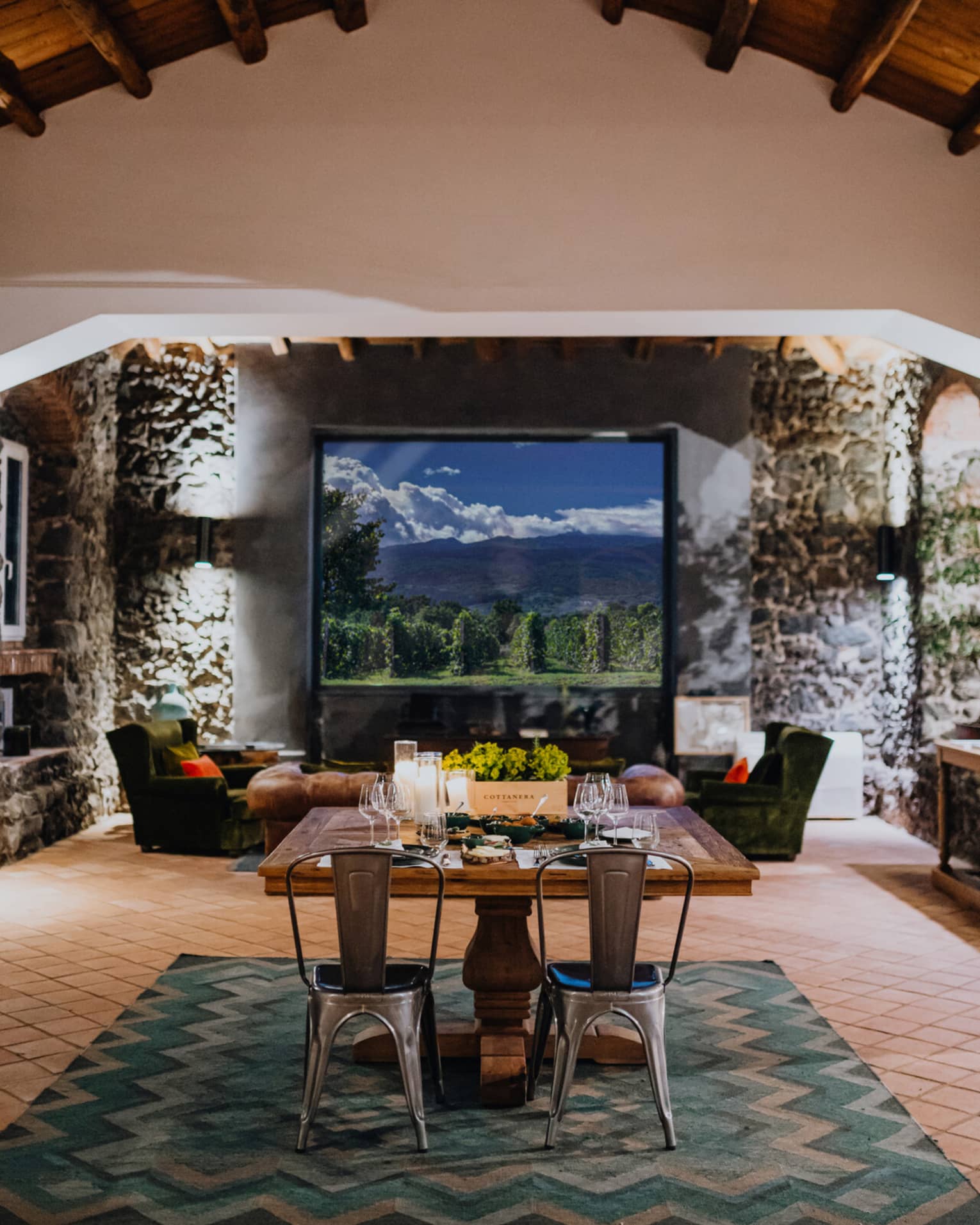 A wooden dining table set with floral arrangements and glassware,  in a room with a window overlooking mountains and a vineyard