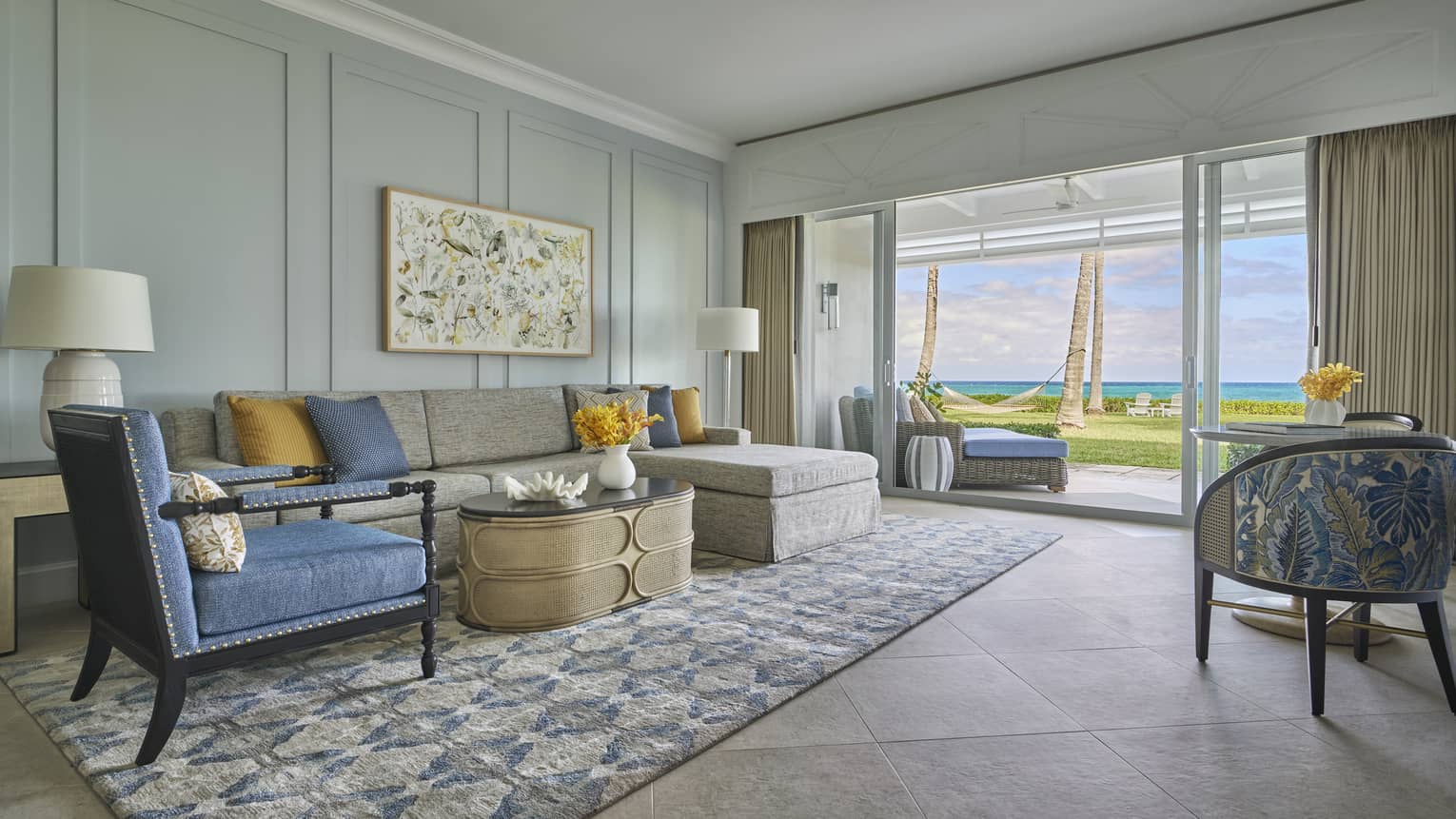 Living room with a gray sectional sofa, blue armchair, patterned rug and glass doors opening to an ocean view with palm trees