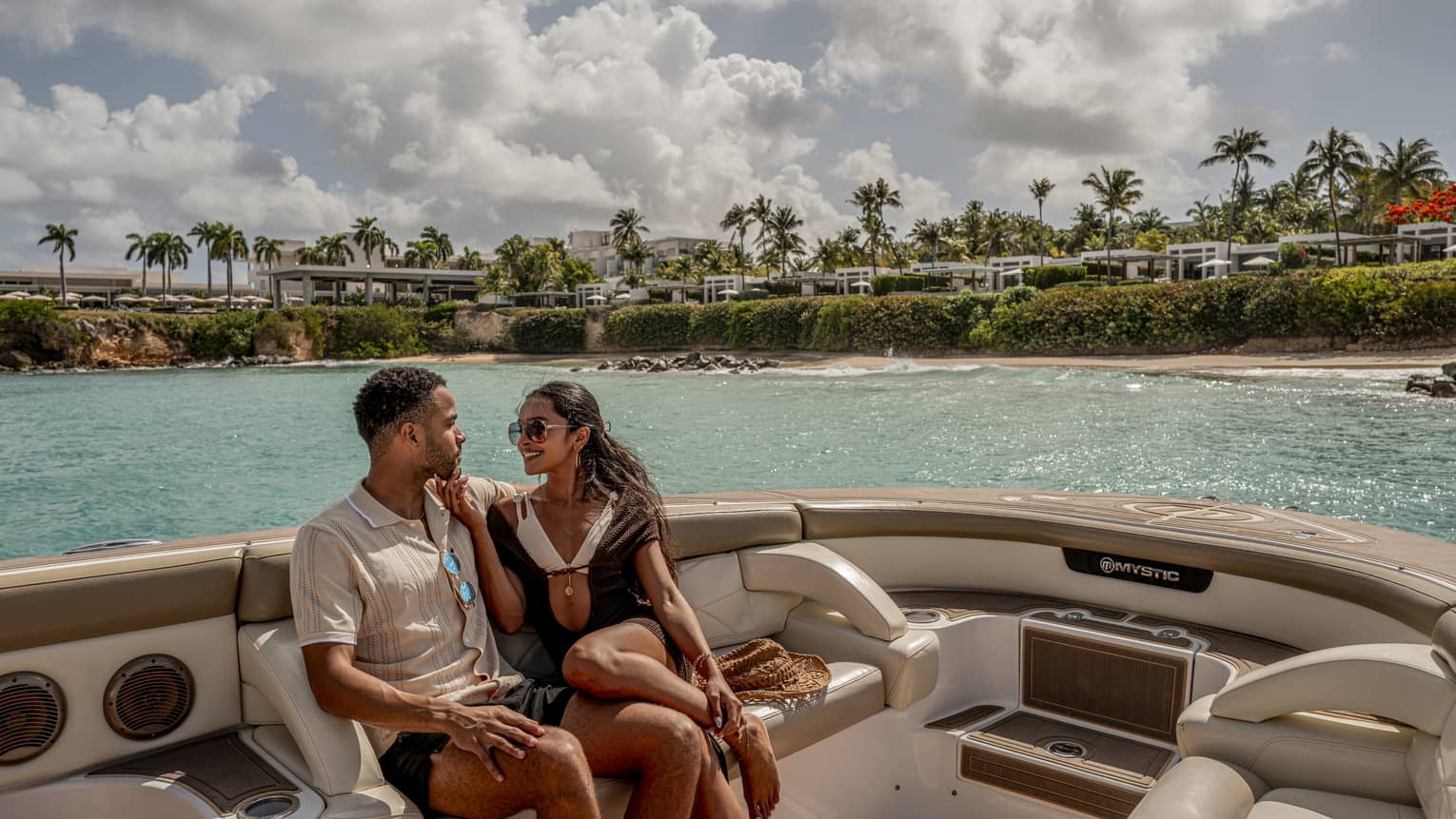 Two people sit on luxury yacht at sea, with palm trees and resort in the background
