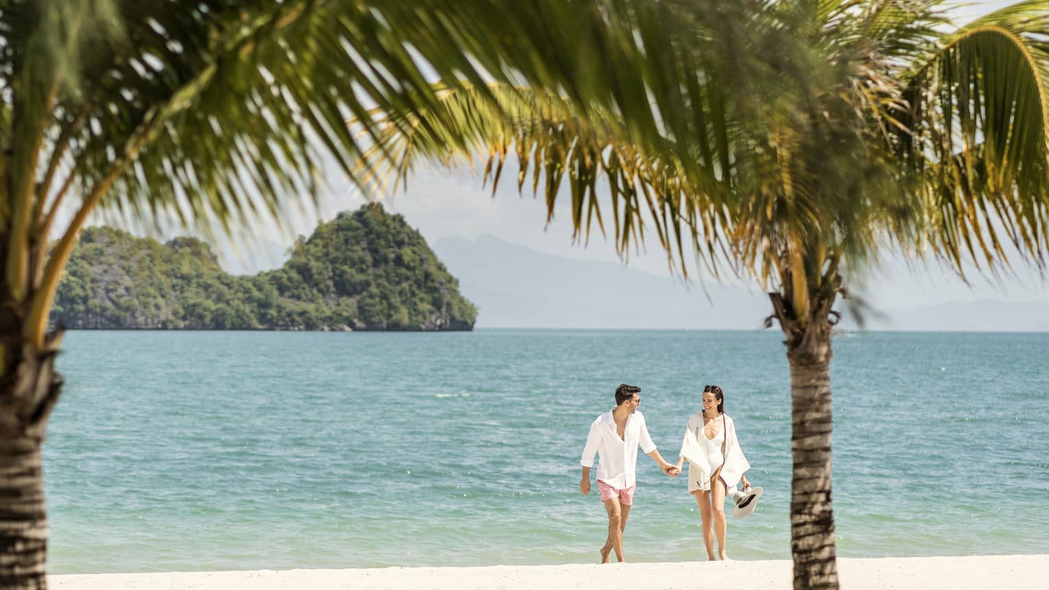 A man and woman dressed in white, holding hands while walking on the beach