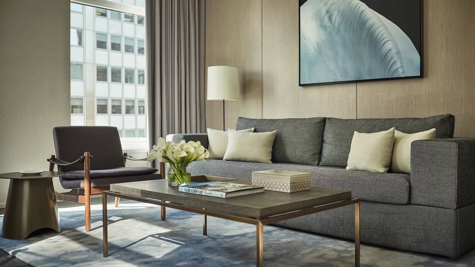 Hudson Suite long grey sofa, table with white flowers, books, armchair by window, building view