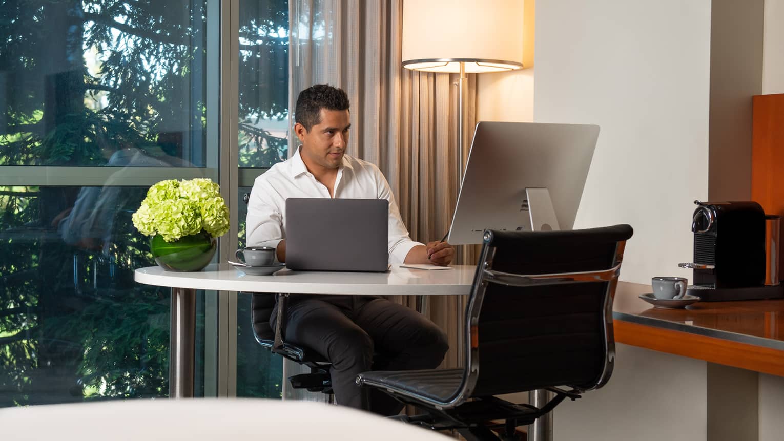Man seated behind white desk with laptop open, extra monitor, flowers in vase, standing lamp