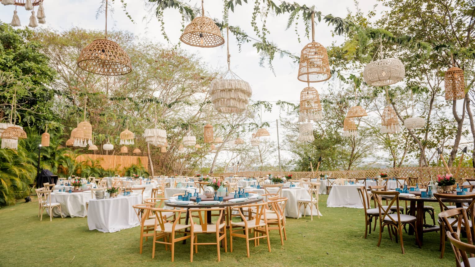 A lawn is set for an outdoor party with various round tables, white table cloths and a variety of straw light fixtures hanging from vined ropes overhead