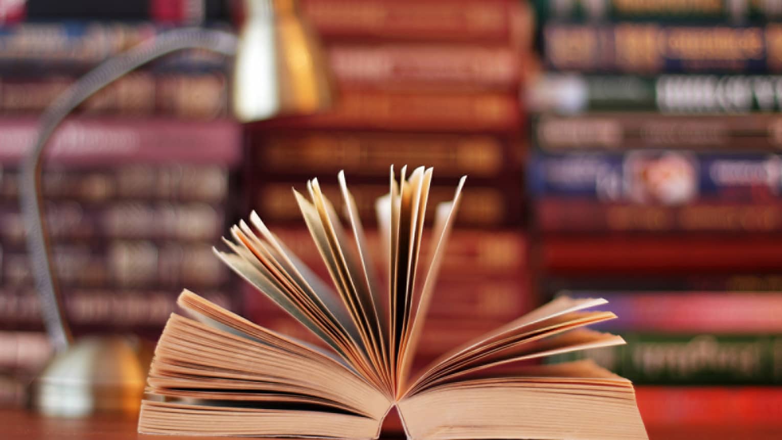 A book is opened, with pages fanned, lamp and stacks of books in background 