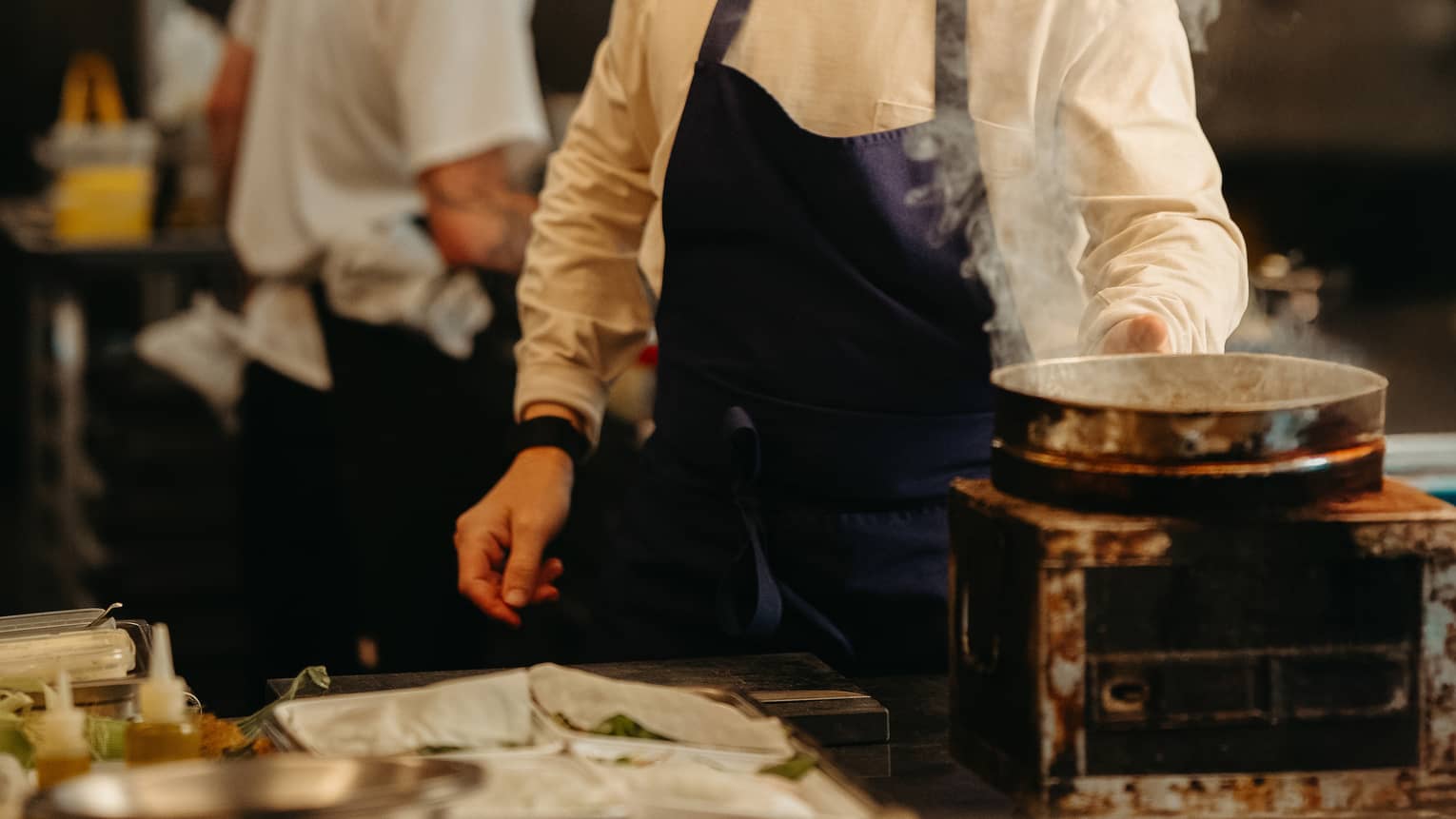 Chef wearing white long-sleeve shirt and dark-blue apron tends to a pot with rising steam