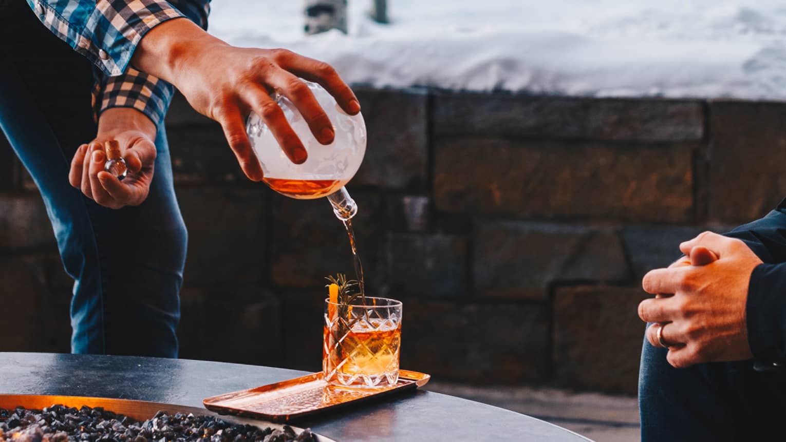 Man in checked shirt pours whisky into cocktail glass by outdoor fireplace, snowy mountain in background