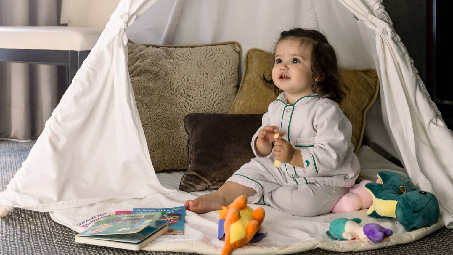 Toddler girl sits among toys and pillows in white play tent in room