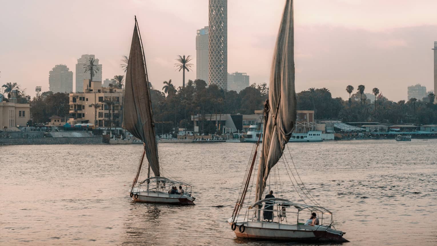 Two felucca boats drift on the water in the evening light, a busy shoreline and Cairo Tower in the distance. 