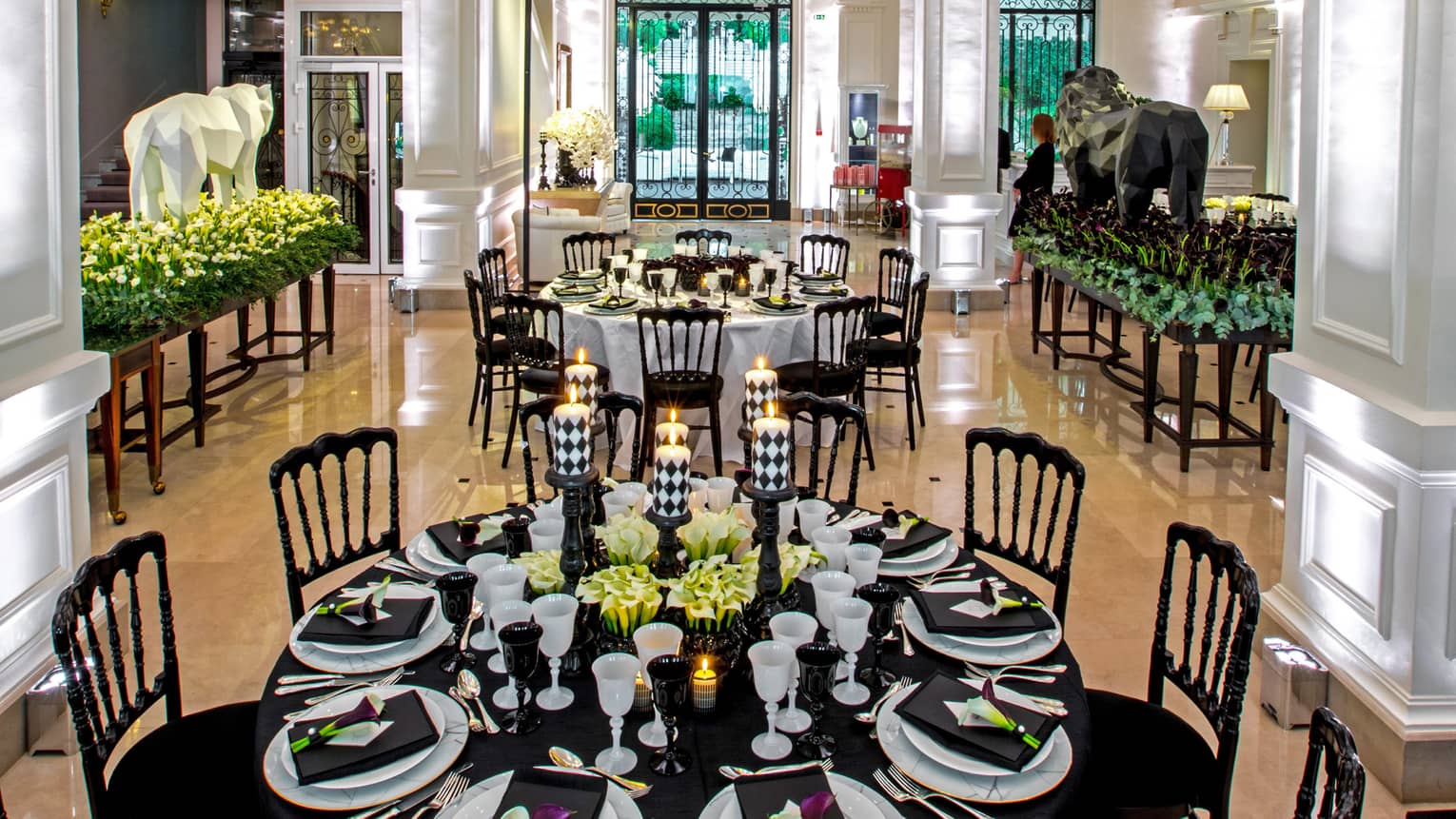 Two elegantly set black round tables, chairs, chandeliers in hotel lobby with white pillars and uplighting