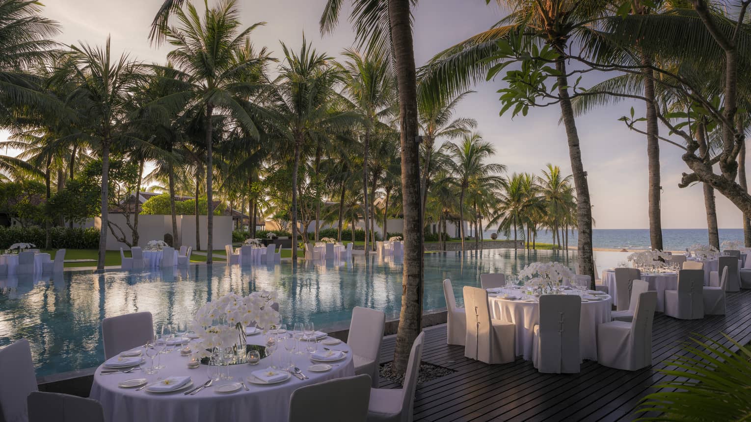 Round dining tables with white tablecloths, white chairs beside infinity pool and palm trees