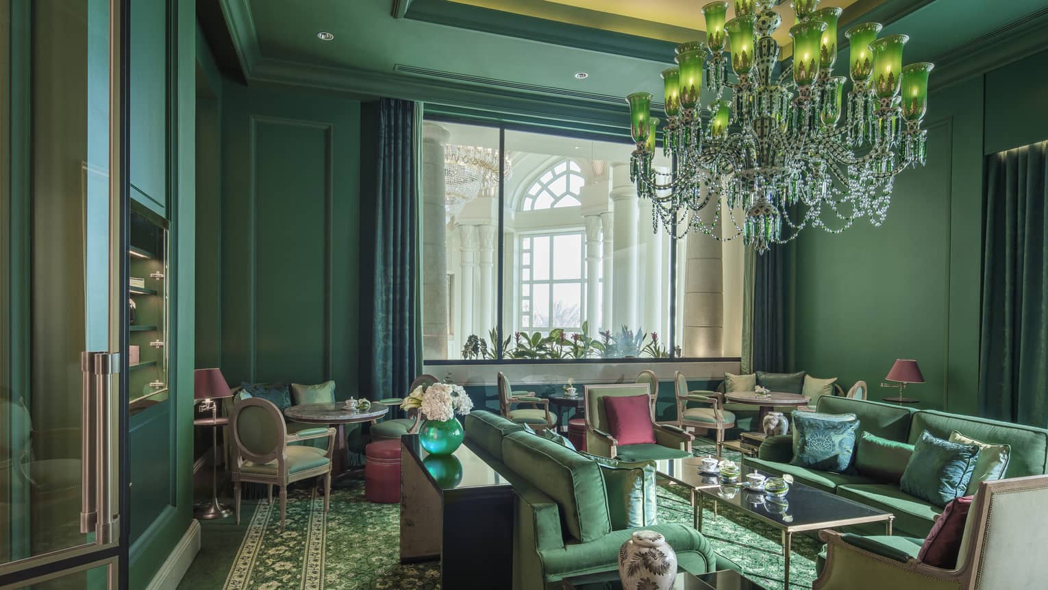 A forest green sitting room with over a dozen velveteen chairs, and a crystal chandelier. 
