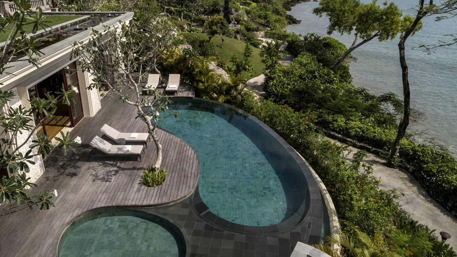 Looking down at Imperial Villa swimming pool and whirlpool on deck with white lounge chairs, garden path and ocean below