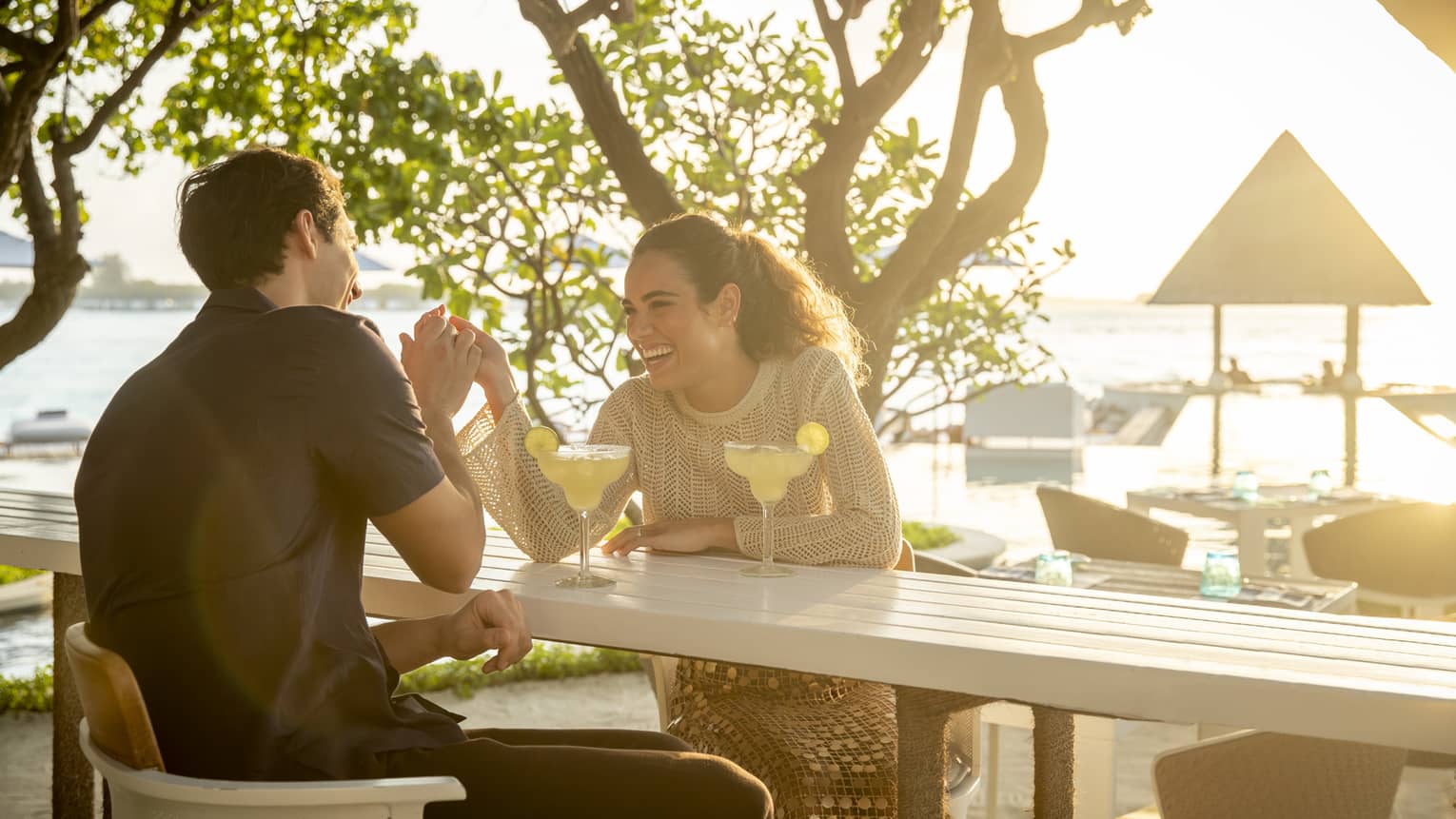 Laughing couple enjoys cocktails at outdoor beachside restaurant surrounded by greenery