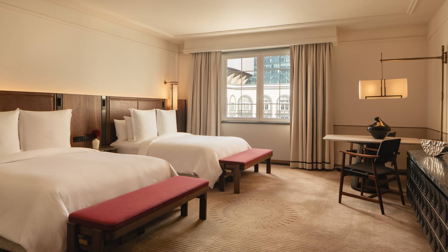 Hotel room with two queen beds, each with a red bench at the foot of the bed, plus a table with two chairs and a window