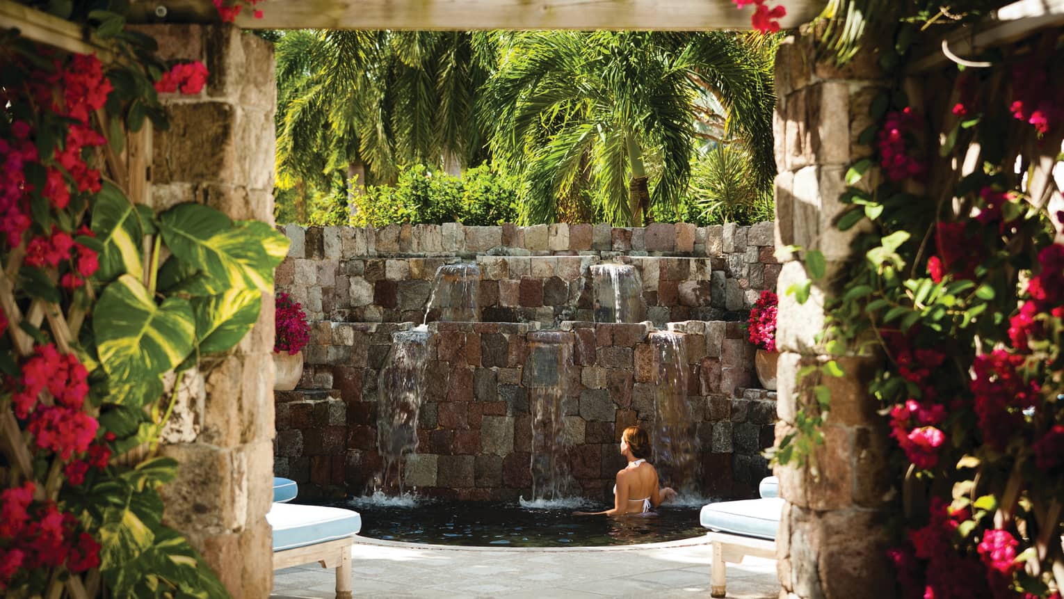Woman bathes in small pool under stone fountain under palm trees, brick walls with pink flowers