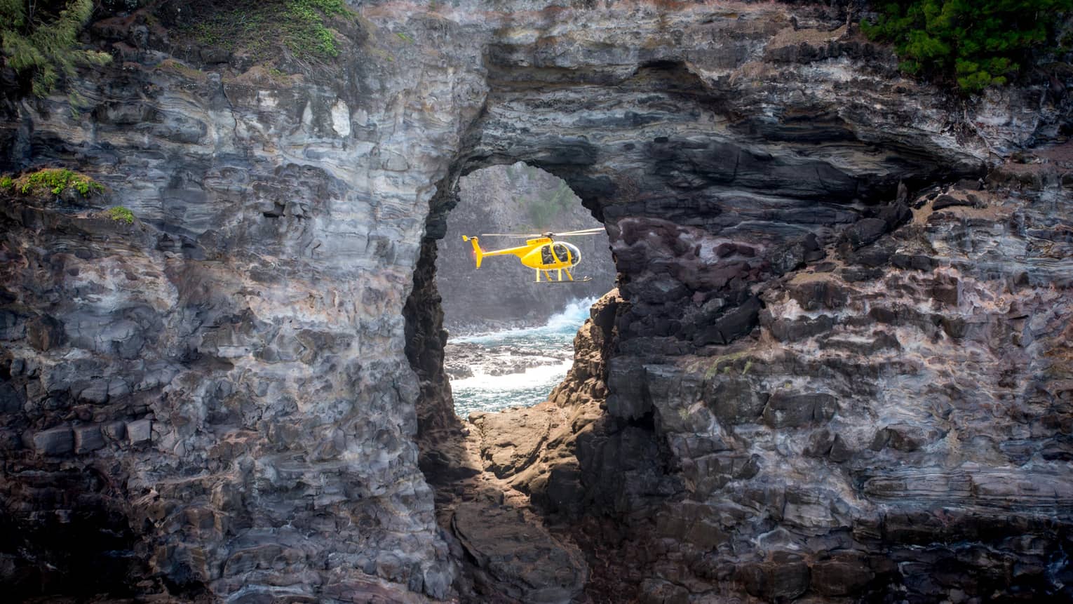 A sideview of a yellow holicopter, seen through an opening in a rock structure. 