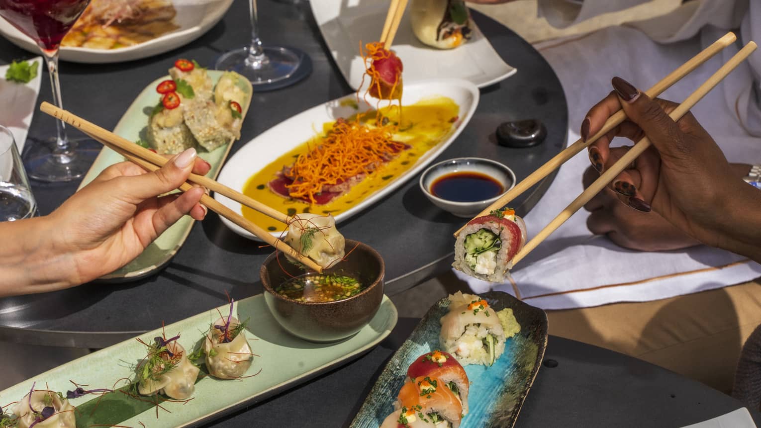 Three hands picking up sushi with chopsticks over plates of dishes