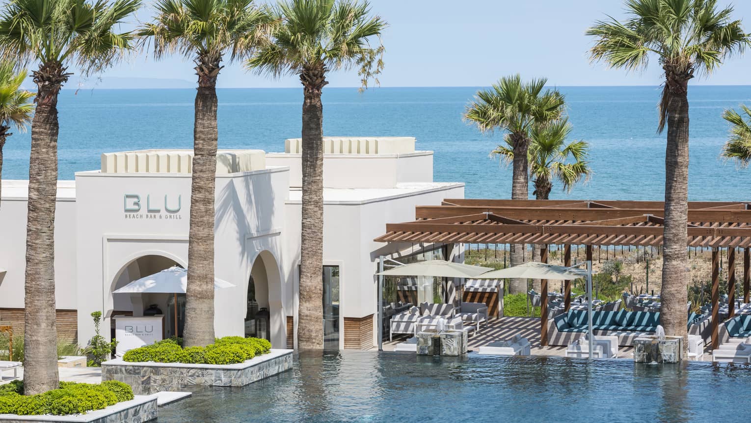 Exterior of BLU Bar & Grill’s white building beside pool surrounded by palm trees, sea in backdrop