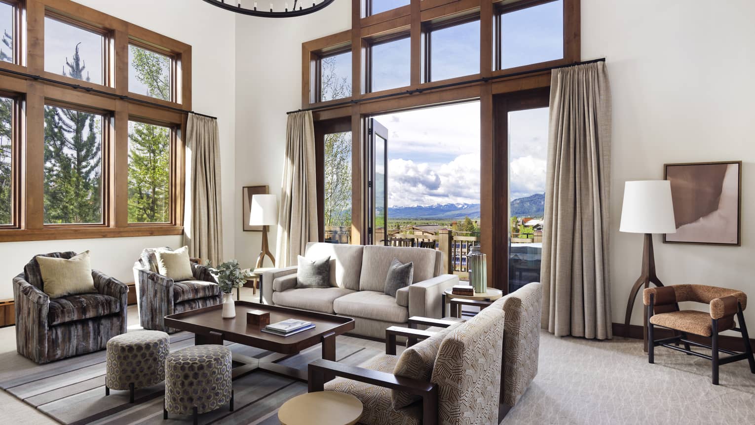 Luxury resort living room with high ceilings, large windows, sofa, armchairs, ottomans, table, floor lamps and doors opening to a mountain-view balcony