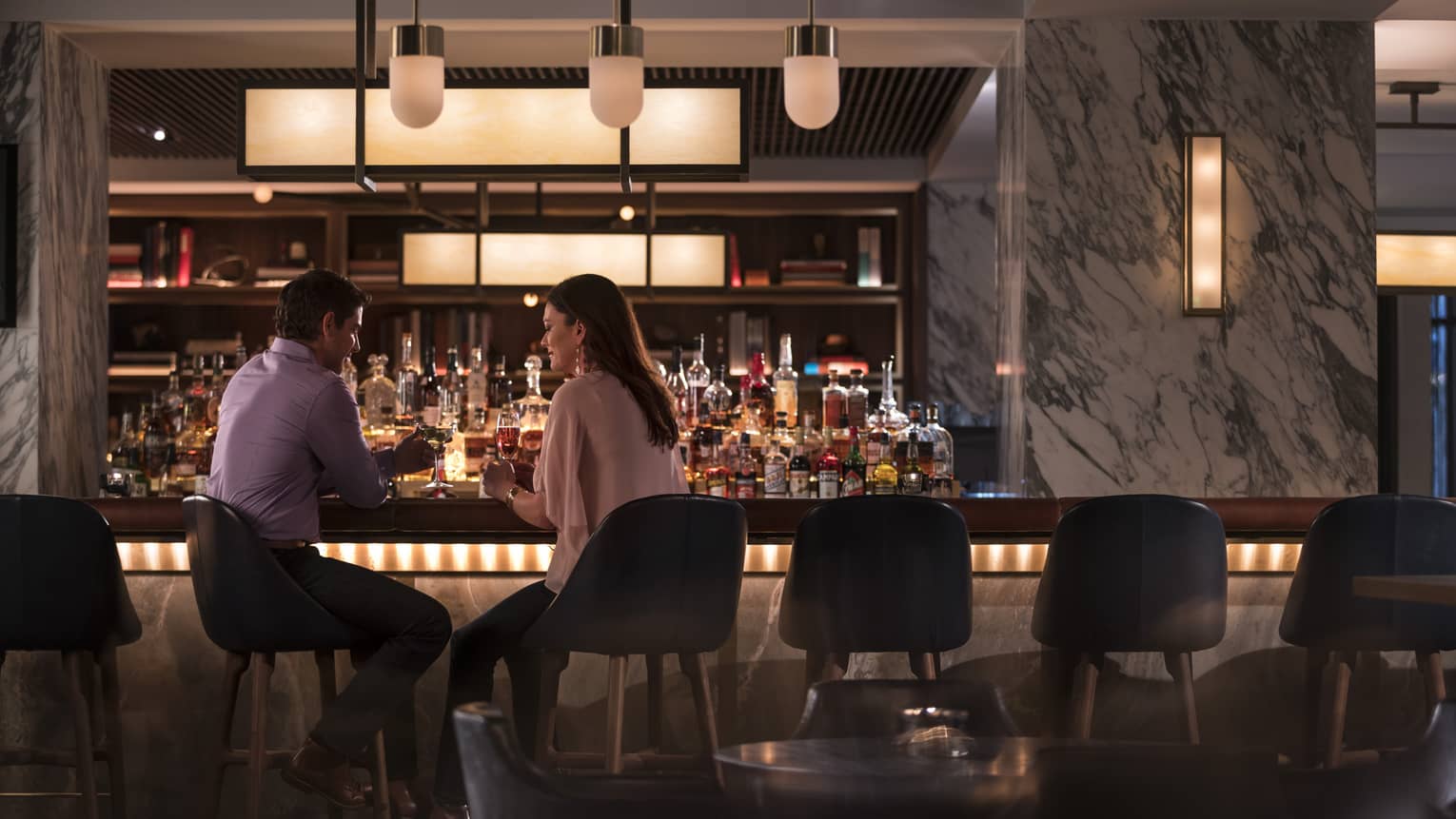 A man and woman sitting at a dimly lit bar with stone walls and a large selection of drinks.