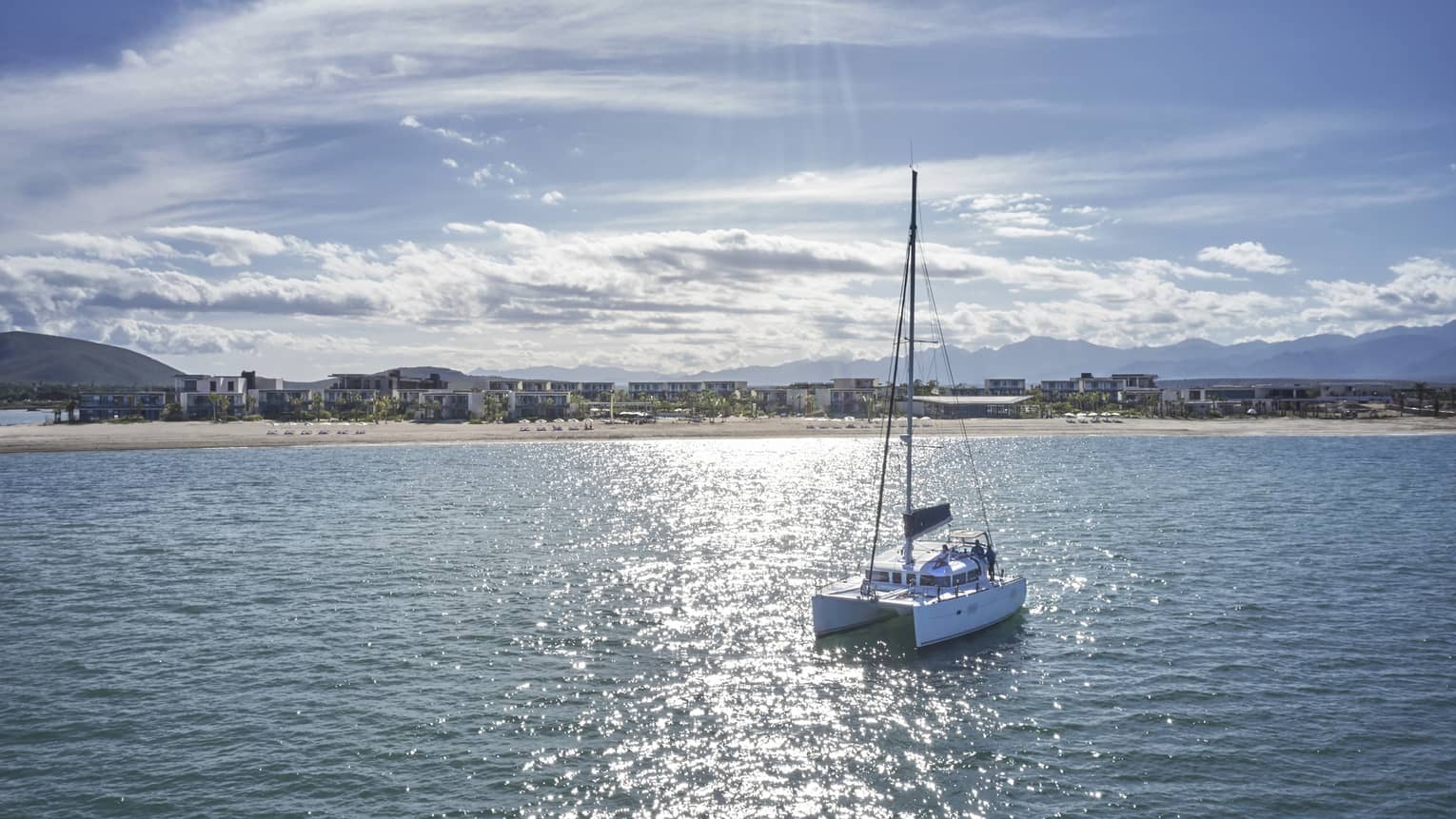 A trimaran boat sails over a glistening sea toward a shore lined with beach homes, distant mountains along the horizon.