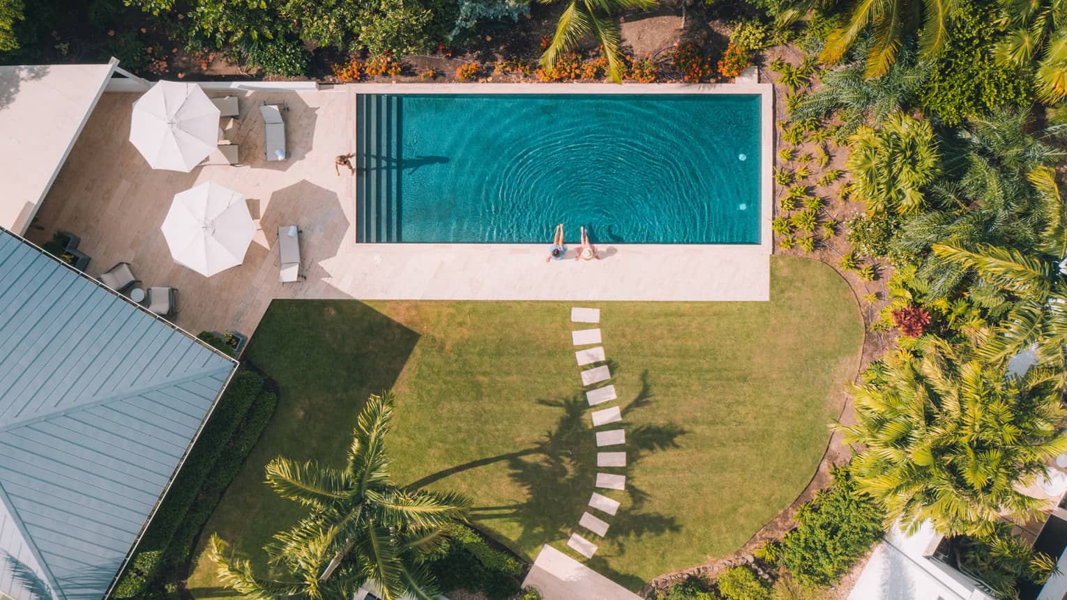 Two people sitting at the edge of a pool surrounded by trees, two umbrellas and a small building.