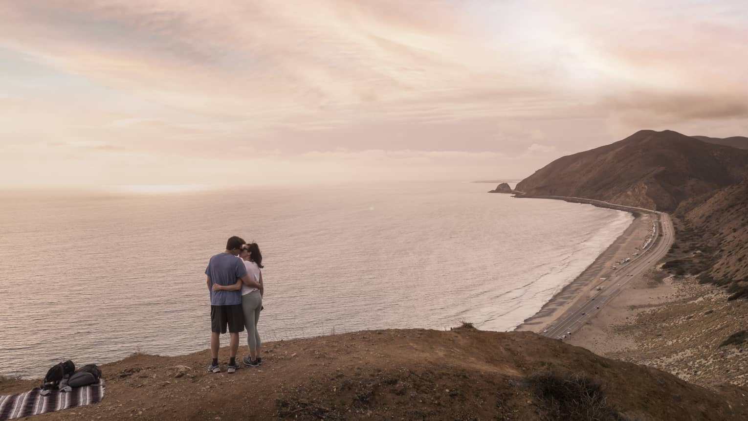 A man and woman hugging while looking out at the ocean on a small hill with a blanket on the grass near them.
