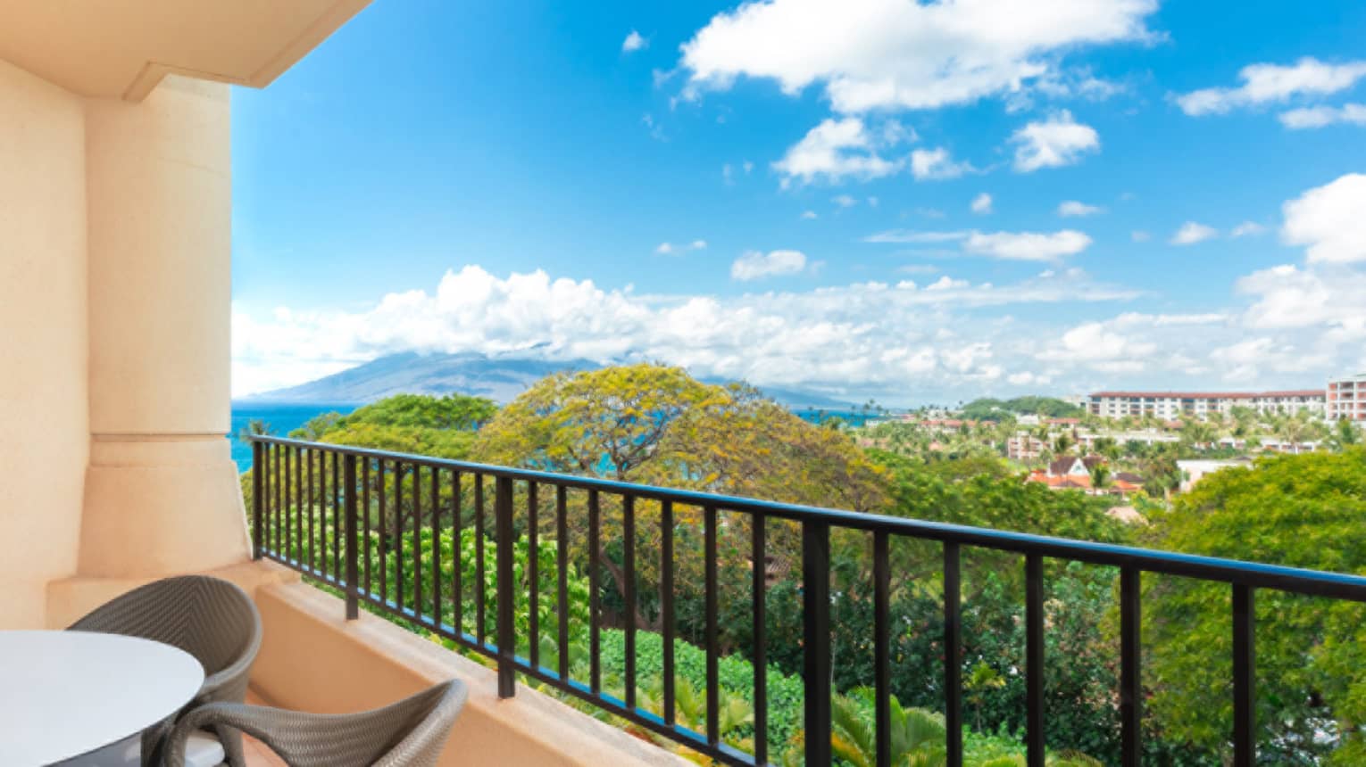 Balcony view overlooking lush greenery, distant buildings and mountain under a bright blue sky with clouds