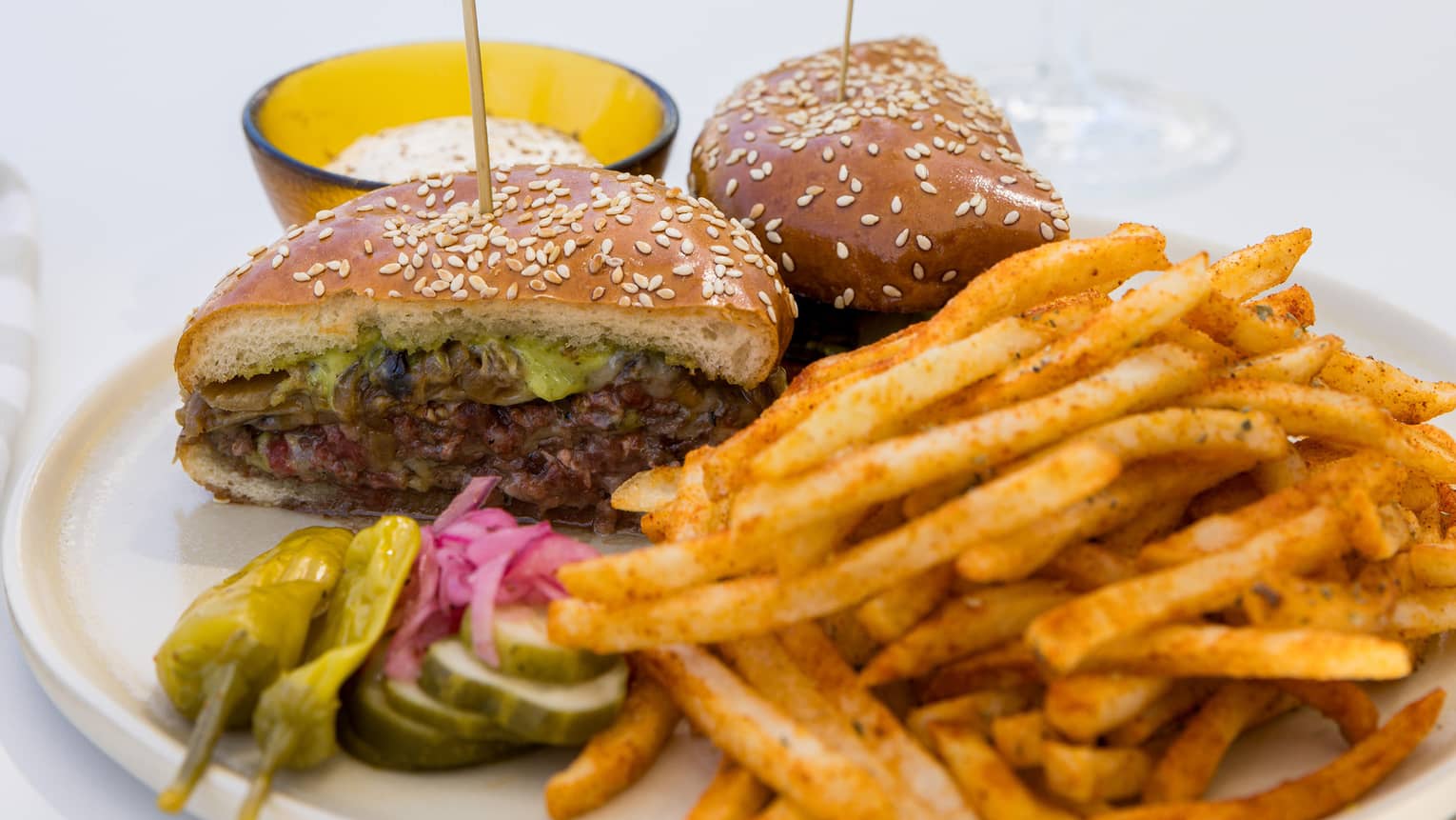Cheeseburger sliced in two served with French fries and garnished with pickled vegetables with a beverage in a tall stemmed glass nearby
