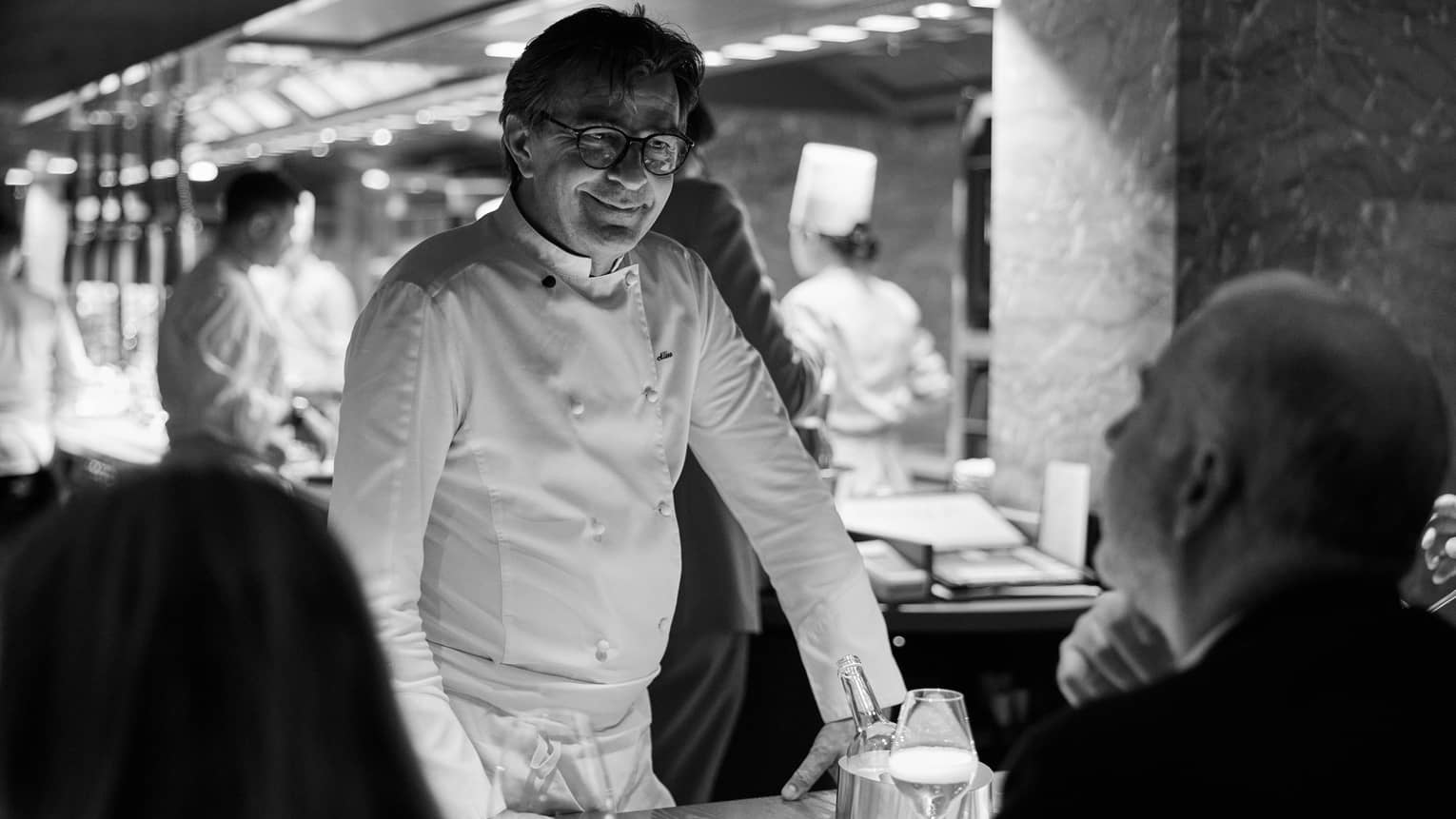 In a restaurant space, a chef stands near a dining table with guests seated at it.