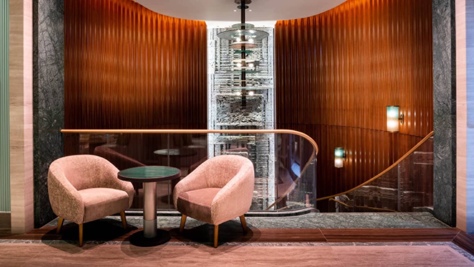 Seating vignette with two rose-coloured club chairs with small round table in centre and beside glass-walled staircase