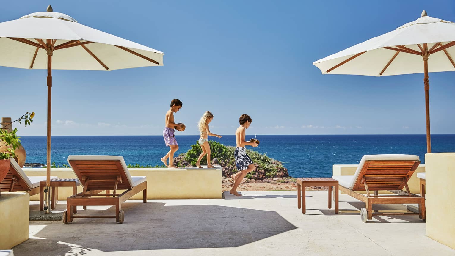 Three children carry coconut drinks along ledge on patio facing ocean