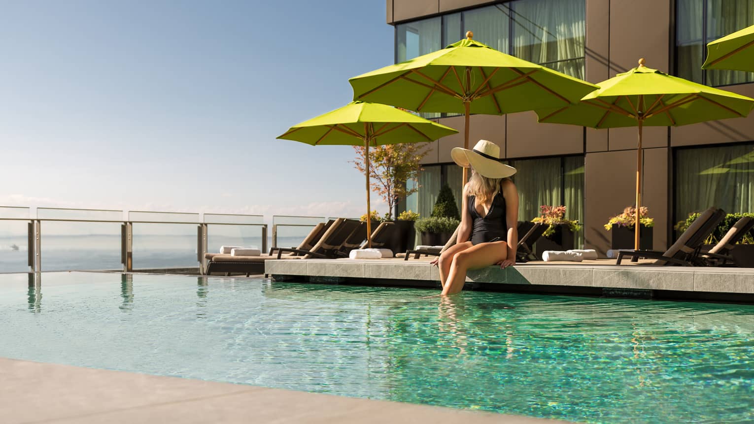 Woman in black swimsuit, sunhat sits on ledge of pool in front of green patio umbrella 