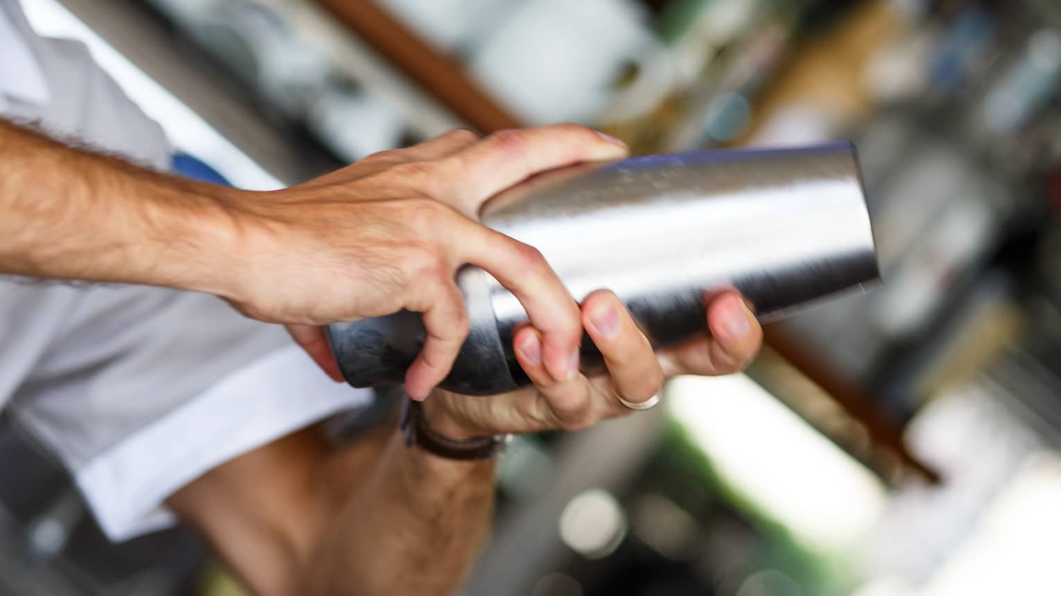 Against a blurred backbar, a bartender’s hands grip a stainless-steel cocktail shaker, a thumb securing the cap in place. 