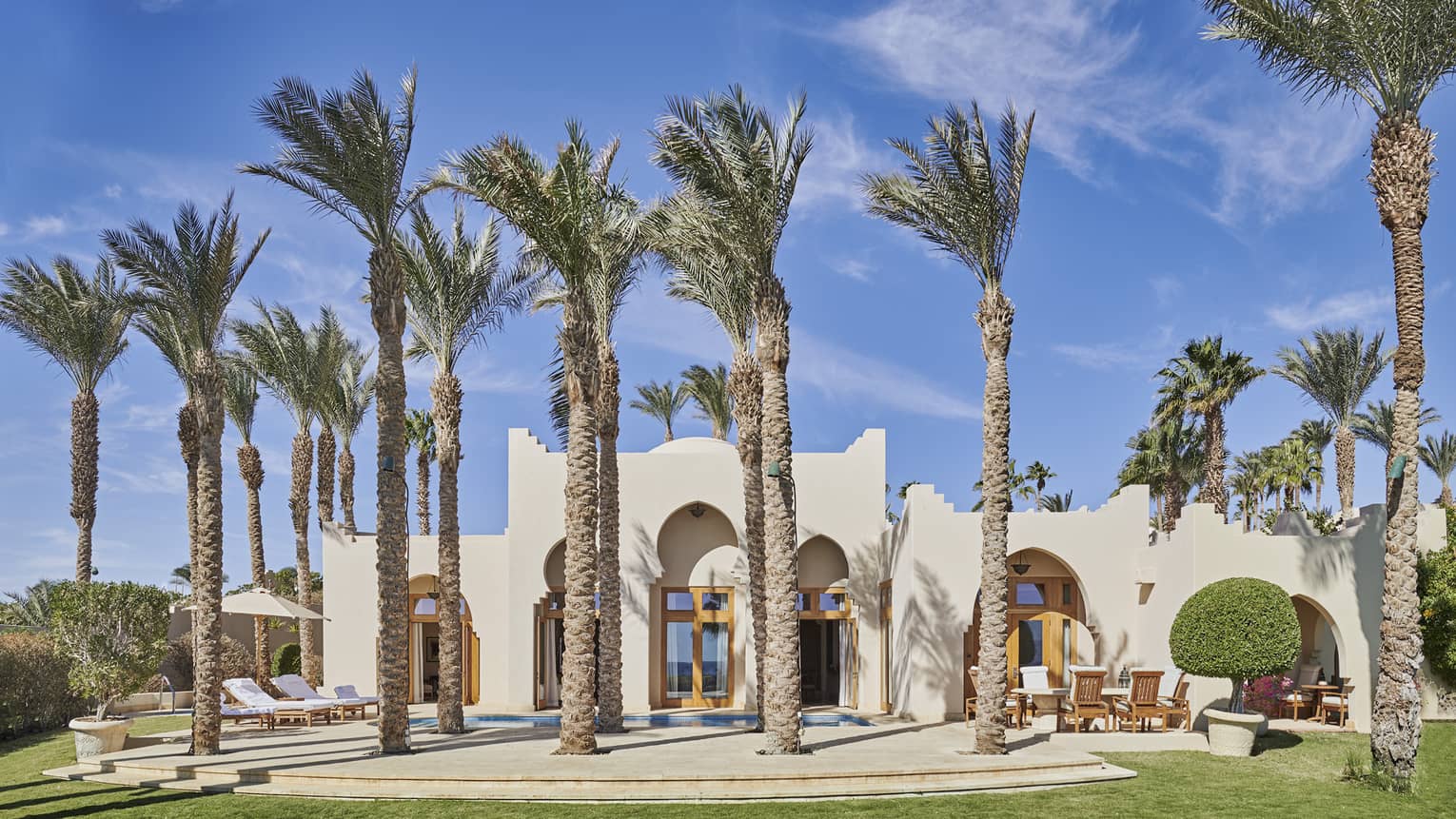 The exterior of a grand villa, with various tall palm trees in front and green grass lawn