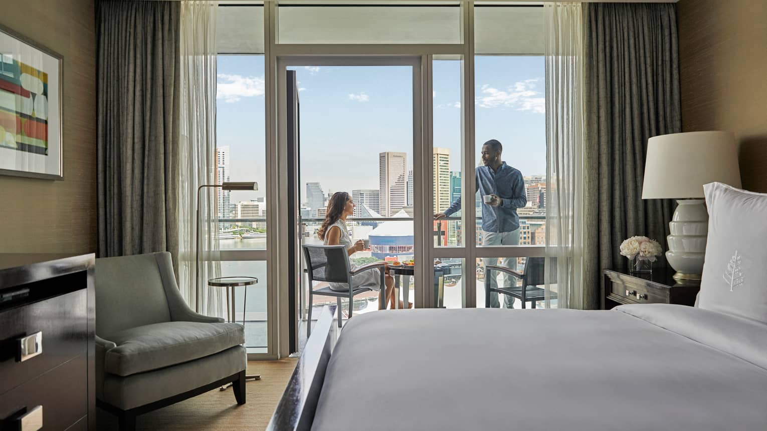 Hotel room with king bed, light grey arm chair, opening to outdoor balcony with two people talking