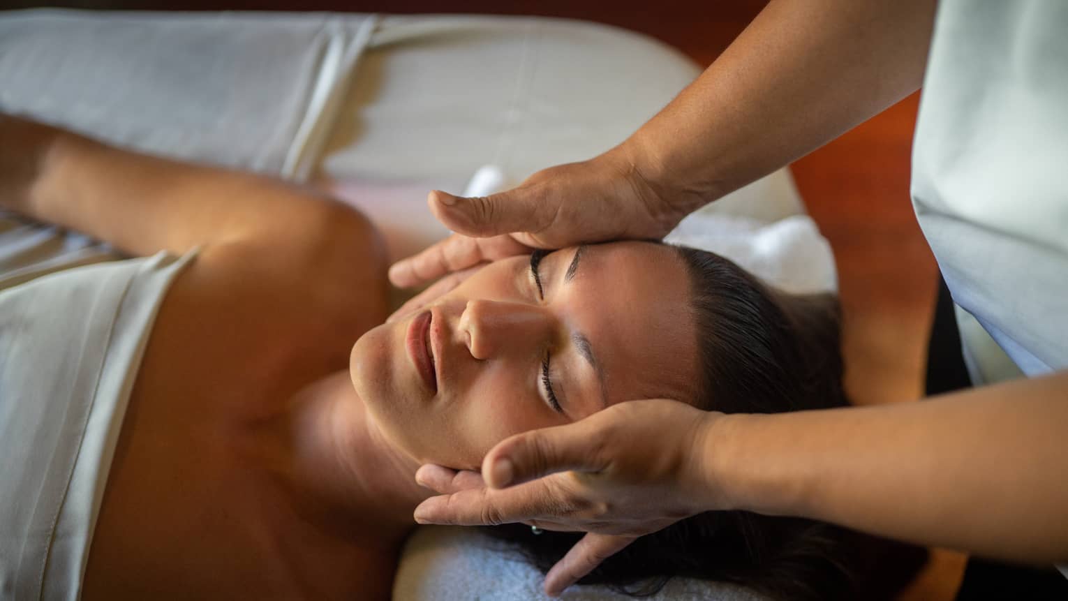 Woman lays on massage table with eyes closed as hands massage her cheeks and chin