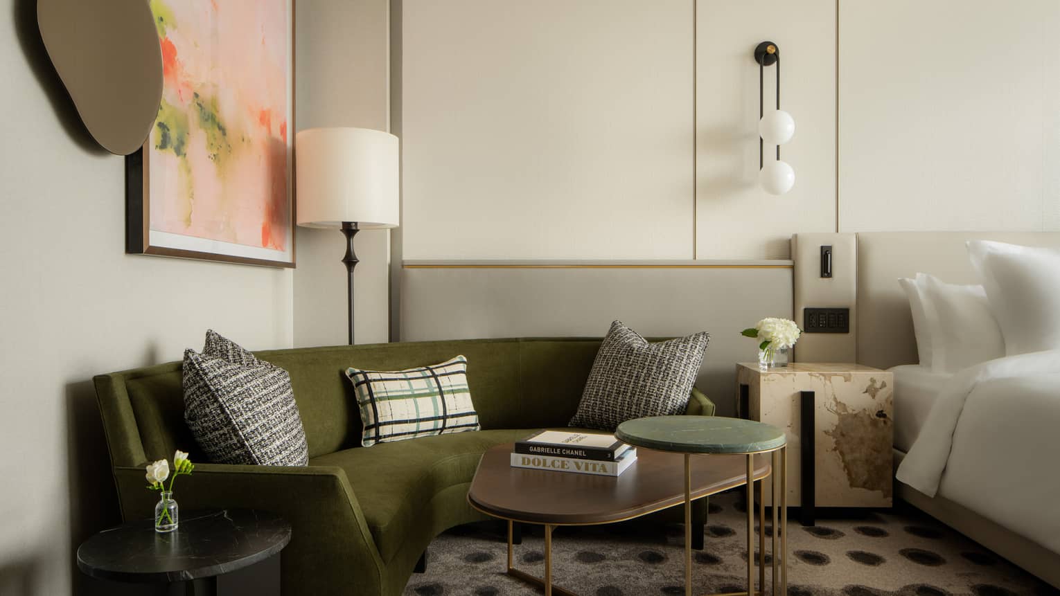Olive green velvet sofa in a suite seating area with a black side table, patterned pillows, and a curved wooden coffee table stacked with designer books