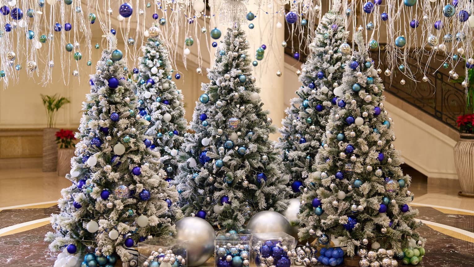 A lobby is decorated with hanging white garland and five frosted Christmas trees adorned with blue and silver ornaments.