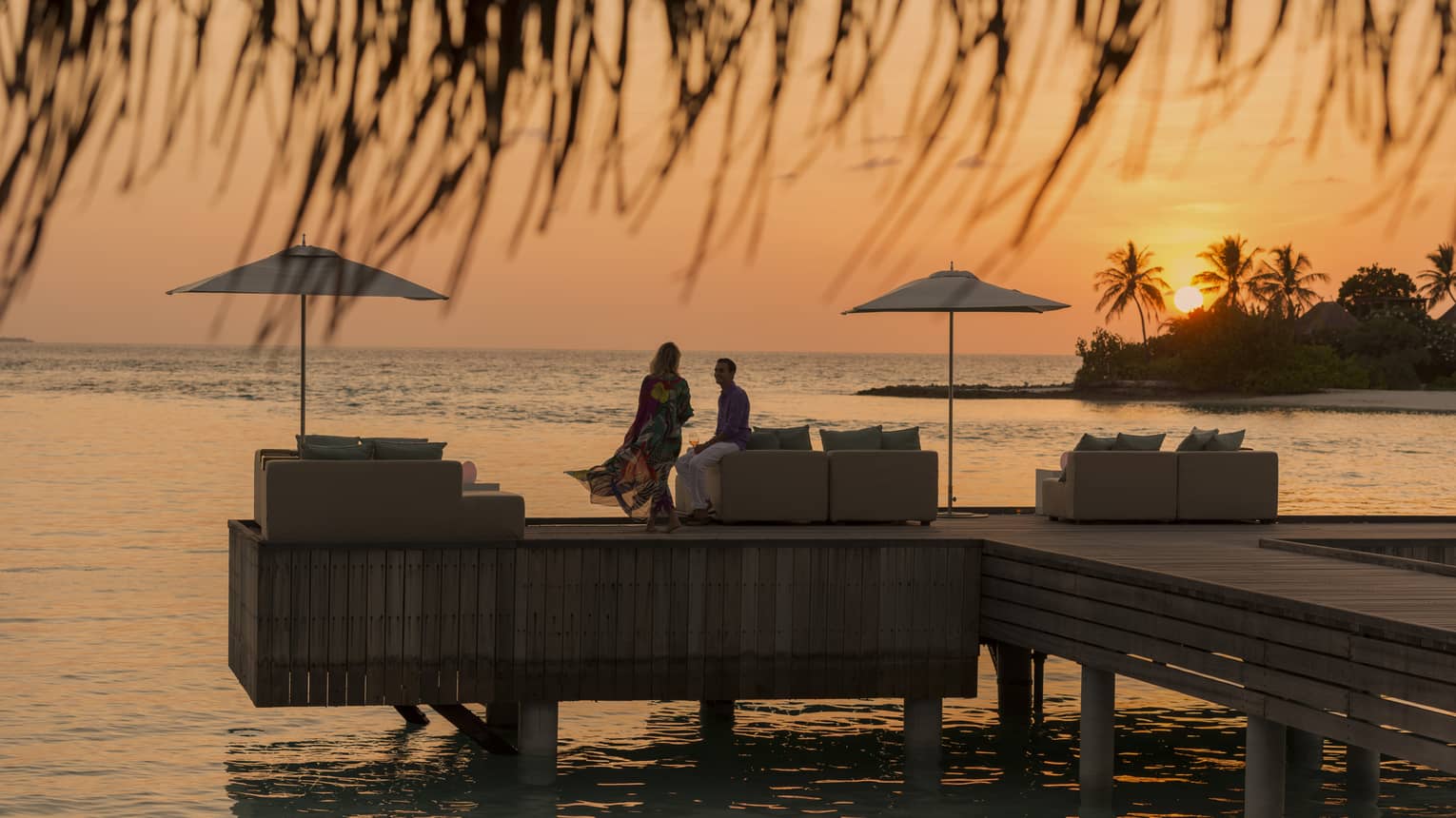 Private island dining at sunset. Woman with long dress blowing in the wind, man sits on the edge of white sofa