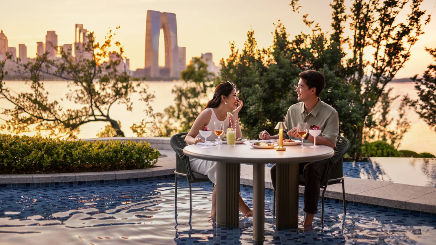Man and woman dine at table set in pool water, with Jinji Lake and Suzhou city centre in the background