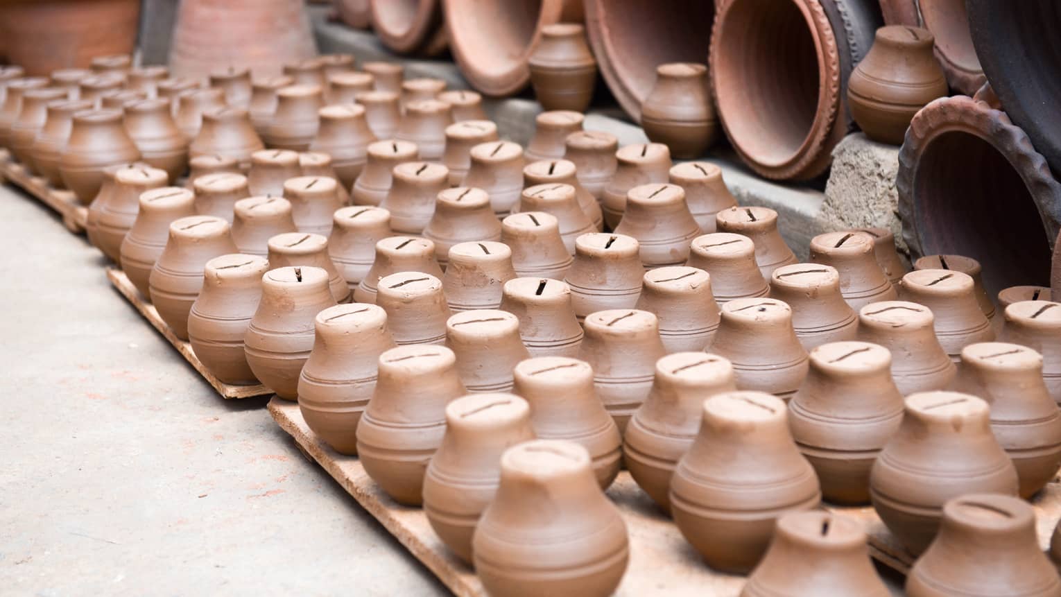 A collection of small, brown clay piggy banks neatly arranged on wooden pallets, surrounded by larger pottery items in the background, creating a display of handcrafted ceramics.