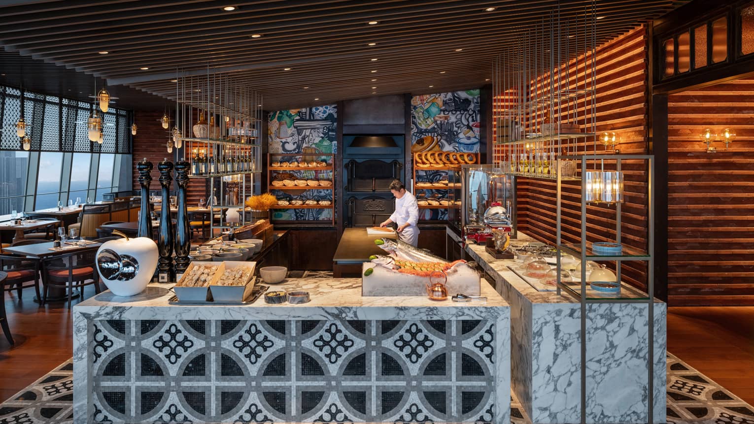 A sushi chef works beside shelves of bread and ornately tiled marble slab counters laden with fresh seafood and delicacies.