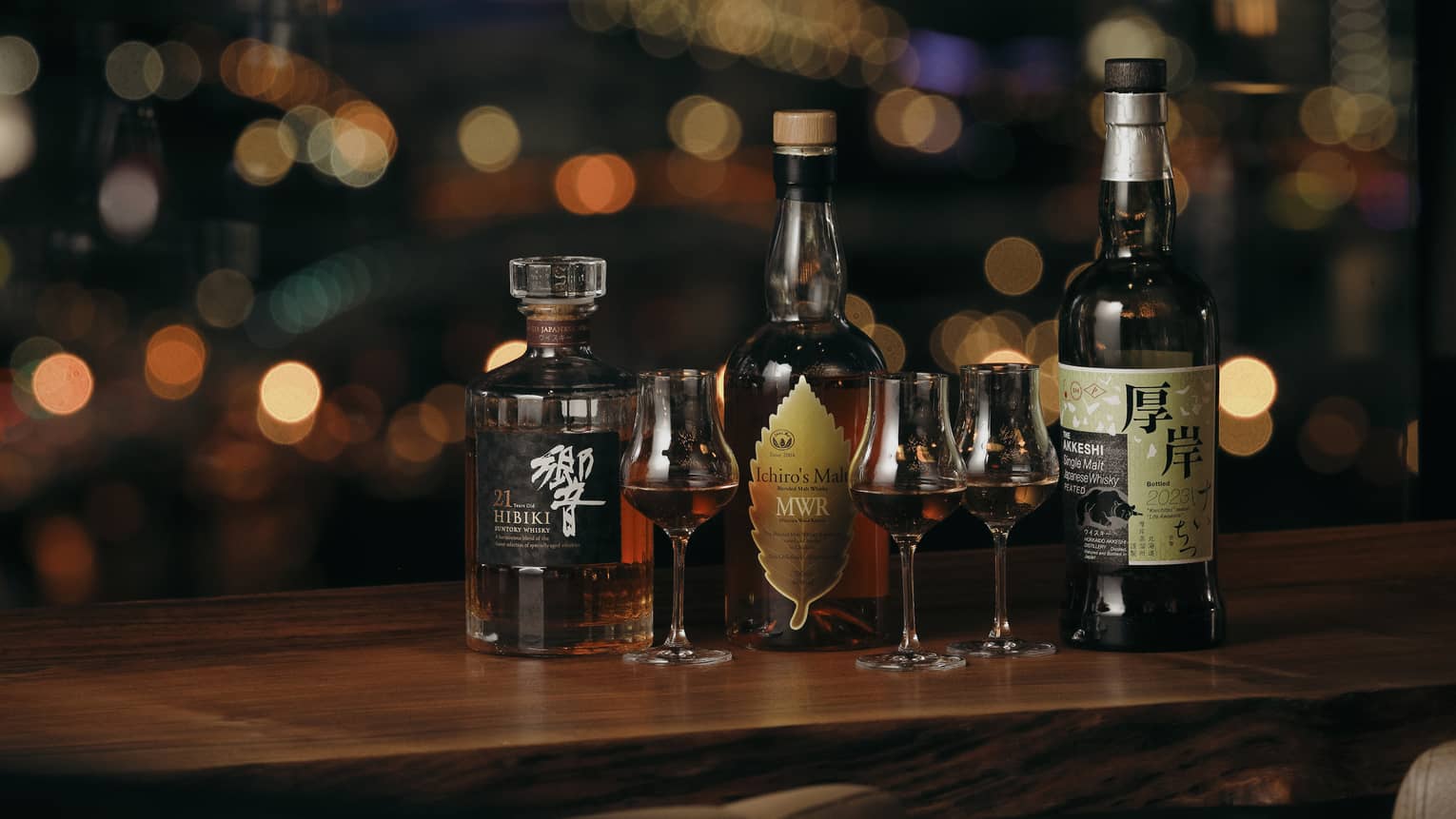 Three Japanese whisky bottles with three stemmed glasses of whisky against a dark background with blurred dots of colour.